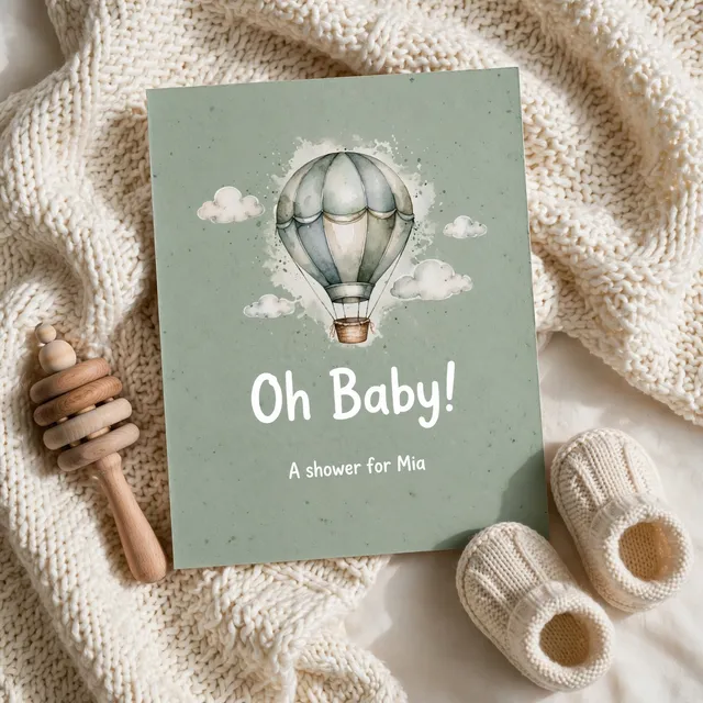 A sage baby shower invitation with a watercolour hot air balloon beside knitted booties.