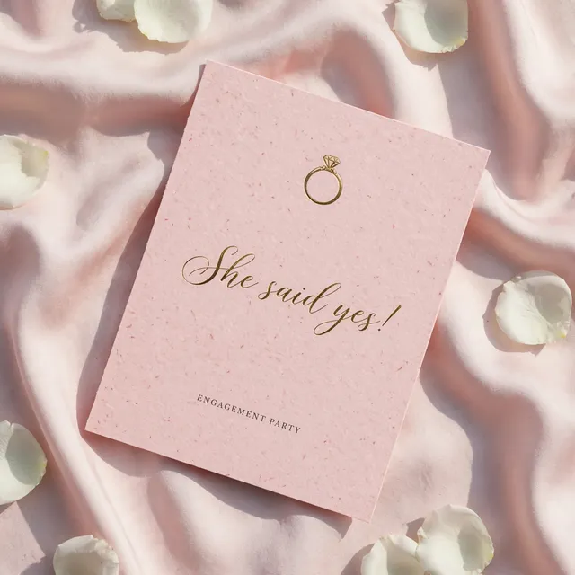 A blush engagement party invitation with a gold ring on pink silk.